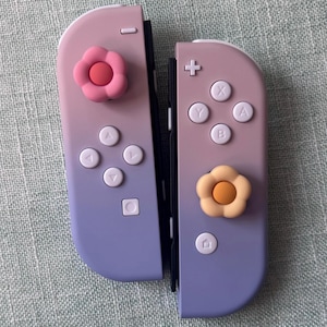 May include: Two game controllers featuring a purple and pink gradient finish. Each controller has white buttons and a silicone joystick cover shaped like a flower, with one pink flower on the left and one peach-colored flower on the right.
