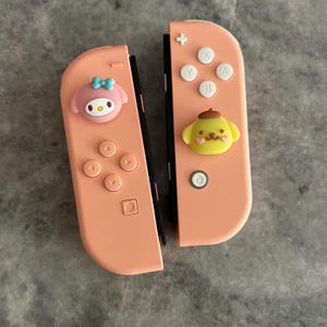 May include: Two coral-colored Nintendo Switch Joy-Con controllers. One features a My Melody character, the other a Pompompurin character. The controllers have buttons with white lettering and symbols.