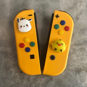 May include: Two yellow Nintendo Switch Joy-Cons with colorful buttons. One Joy-Con features a white bunny face, and the other has a yellow chick face. The controllers are designed for gaming.
