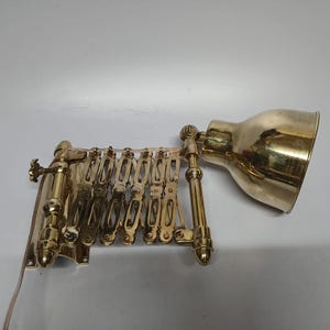 May include: A brass-colored, articulated wall-mounted lamp. The lamp features a concertina-style arm and a bell-shaped shade. The lamp is designed to be adjustable and is mounted on a wall plate. The lamp has a vintage aesthetic.