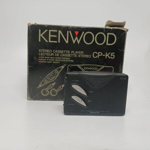 以下が含まれることがあります： 黒いKenwood CP-K5ステレオカセットプレーヤーとオリジナルの箱。箱は黒で、白い文字と赤い三角形のロゴが付いています。プレーヤーにはシルバーのボタンが付いています。