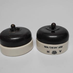 以下が含まれることがあります： ダークブラウンのトップとクリーム色のベースを備えた2つの丸い電気トグルスイッチ。右側のスイッチのクリーム色のベース側面には、REF. No 5C/622というテキスト、文字AとM、および小さな王冠のシンボルが印刷されています。