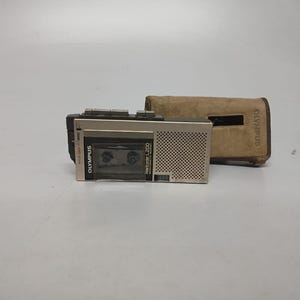 May include: A vintage Olympus Pearlcorder L200 microcassette recorder in silver and black. It is next to a brown leather case. The recorder has the Olympus logo and model details printed on it. A retro audio device.