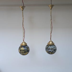 May include: Two pendant lights with globe-shaped glass shades. The shades feature a mix of blue, yellow, and black colors. Each light has a brass-colored fixture and chain, suspended from the ceiling.