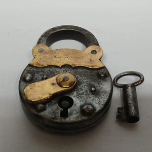 May include: A vintage round padlock with a weathered dark metal body and a brass plate featuring the text MADE IN GERMANY. The lock includes a brass keyhole cover and a matching small metal key positioned to the right of the main lock body.