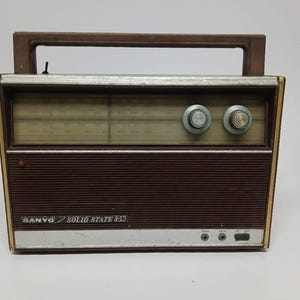 Puede incluir: Radio vintage Sanyo Solid State 852. La radio tiene un panel frontal marrón con un borde plateado y dos perillas plateadas. La parte superior tiene un asa marrón. El texto "SANYO SOLID STATE 852" es visible.