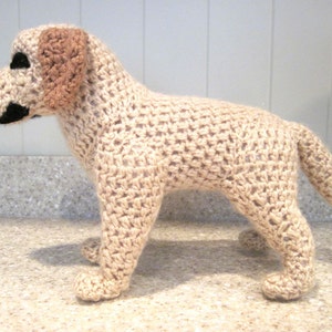 Op de afbeelding: Een gehaakt hondenspeelgoed in een lichtbeige kleur met bruine oren. De hond staat op alle vier poten met zijn staart opgerold.