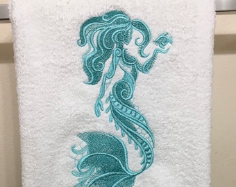 May include: White bath towel with a teal embroidered mermaid design. The mermaid is holding a small fish in her hand.