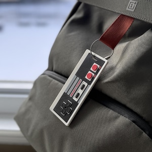 May include: A gray backpack with a red and white Nintendo controller keychain. The keychain has the words "SELECT START" printed on it.