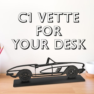 May include: Black metal model of a C1 Corvette on a black base. The model is on a wooden table with pencils and a cup in the background. The text "C1 VETTE FOR YOUR DESK" is on the wall behind the model.
