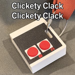 May include: A white and black controller keychain with two red buttons. The keychain has the text "Clickety Clack Clickety Clack" on it.