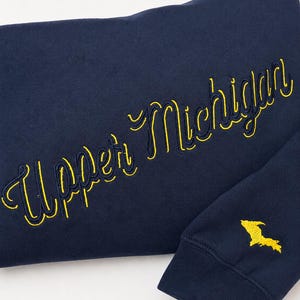 Peut inclure: Un sweat-shirt bleu marine avec le texte Upper Michigan brodé en lettres cursives jaunes sur la poitrine, accompagné d'une petite carte brodée en jaune de la péninsule supérieure du Michigan sur le poignet côtelé de la manche.