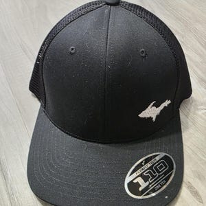 Può includere: Un cappellino da baseball nero con retro in rete e pannello frontale solido. Il cappellino presenta un contorno ricamato in bianco della penisola superiore del Michigan sul davanti. Un adesivo circolare sulla visiera riporta la scritta Flexfit Tech 110 One Ten.