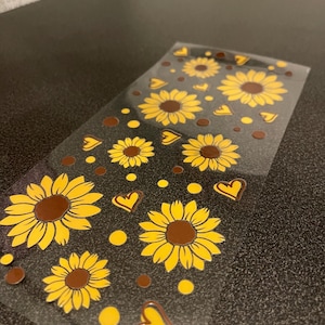 Könnte beinhalten: Transparente Aufkleberbogen mit gelben Sonnenblumen mit braunen Zentren, gelben Herzen und gelben Punkten auf dunklem Hintergrund. Die Aufkleber sind in einem sich wiederholenden Muster angeordnet. Das Blatt liegt auf einer dunklen Oberfläche.