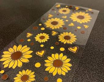 May include: Clear sticker sheet featuring yellow sunflowers with brown centers, yellow hearts, and yellow dots on a dark background. The stickers are arranged in a repeating pattern. The sheet is on a dark surface.