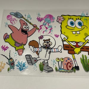 May include: A clear sticker sheet featuring cartoon characters from the animated series SpongeBob SquarePants. The characters include SpongeBob, Patrick Star, Sandy Cheeks, Plankton, and Gary the snail, along with jellyfish and coral.