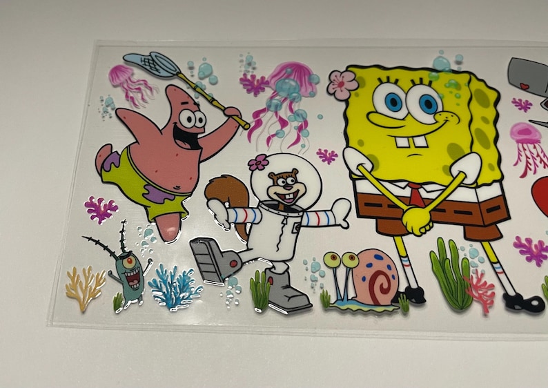 May include: A clear sticker sheet featuring cartoon characters from the animated series SpongeBob SquarePants. The characters include SpongeBob, Patrick Star, Sandy Cheeks, Plankton, and Gary the snail, along with jellyfish and coral.
