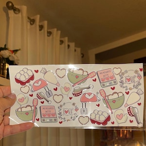 May include: A sheet of stickers with a baking theme. The stickers feature various kitchen tools, including a mixer, measuring cups, a rolling pin, and a piping bag. The stickers are decorated with hearts and the words "Measured with Love" and "Made with Love".