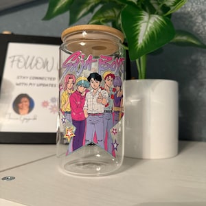 Puede incluir: Un vaso de vidrio transparente con tapa de bamb&uacute;, con un gr&aacute;fico colorido de seis personajes de estilo anime y el texto "Saja Boys". El dise&ntilde;o incluye estrellas y una paleta de colores pastel. El vaso se muestra sobre una superficie blanca.