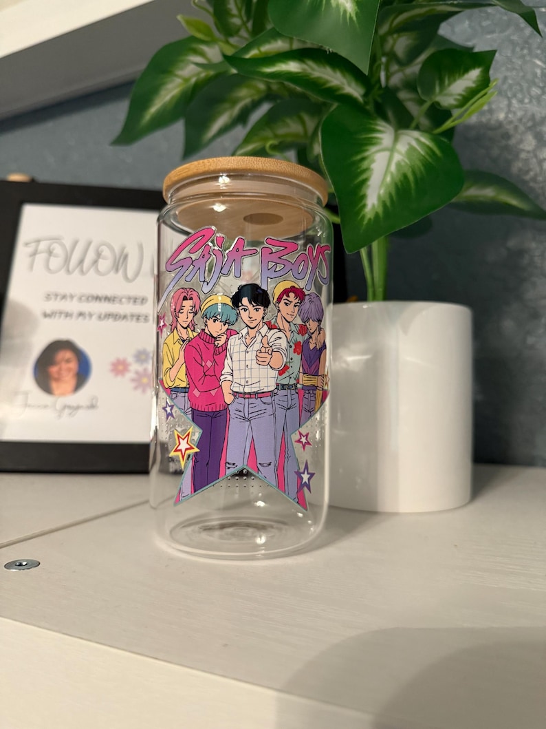 Puede incluir: Un vaso de vidrio transparente con tapa de bamb&uacute;, con un gr&aacute;fico colorido de seis personajes de estilo anime y el texto "Saja Boys". El dise&ntilde;o incluye estrellas y una paleta de colores pastel. El vaso se muestra sobre una superficie blanca.
