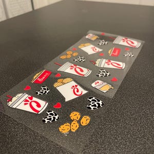 May include: A clear sheet of stickers featuring various Chick-fil-A themed designs. The stickers include a chicken sandwich, waffle fries, a milkshake, a chicken nugget, and a heart with a cow print. The stickers are perfect for adding a touch of Chick-fil-A fun to your belongings.