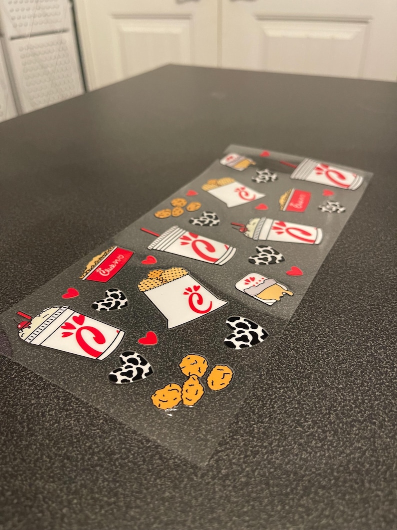 Clear vinyl sticker sheet featuring a fast food-themed design inspired by a popular chicken restaurant. The sheet includes illustrations of cups with red straws, waffle fries, cookies, chicken nuggets, and sauce packets, along with hearts and cow-print patterned hearts. The items are brightly colored in red, white, black, and tan, arranged playfully on a transparent background,