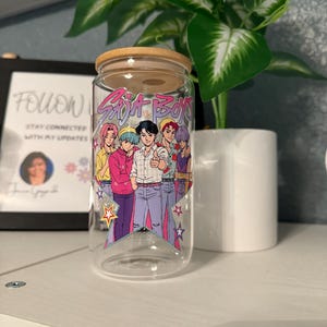 Puede incluir: Un vaso de vidrio transparente con tapa de bamb&uacute;, con un gr&aacute;fico colorido de seis personajes de estilo anime y el texto "Soja Boys". El dise&ntilde;o incluye estrellas y una base triangular. El vaso est&aacute; sobre una estanter&iacute;a blanca.