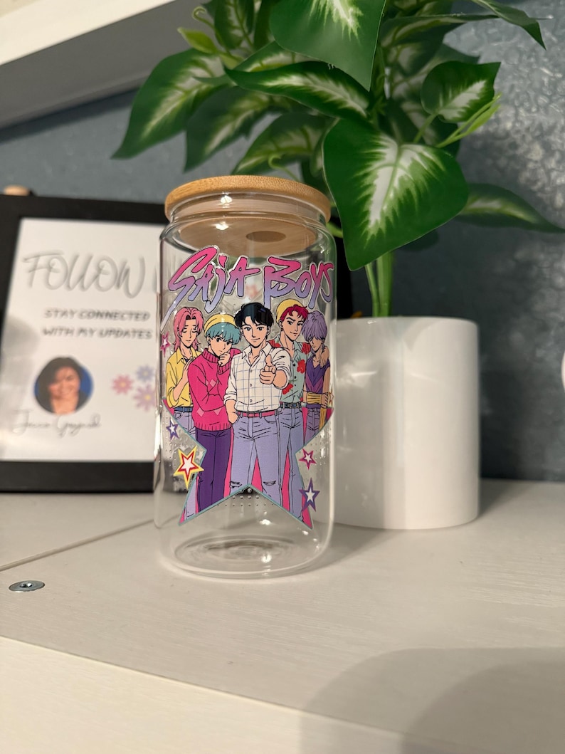 Puede incluir: Un vaso de vidrio transparente con tapa de bamb&uacute;, con un gr&aacute;fico colorido de seis personajes de estilo anime y el texto "Soja Boys". El dise&ntilde;o incluye estrellas y una base triangular. El vaso est&aacute; sobre una estanter&iacute;a blanca.