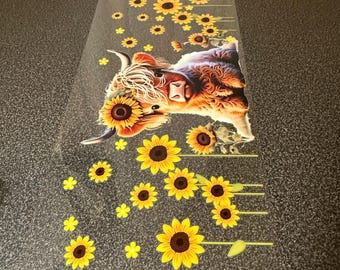 May include: A clear, rectangular sticker featuring a Highland cow with a sunflower crown and sunflowers. The cow has brown and white fur, with yellow and brown sunflowers. Additional yellow sunflowers are scattered around the cow.