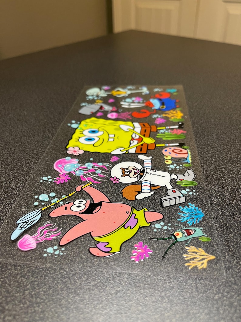 May include: A clear sticker sheet featuring cartoon characters from the animated series. The sheet includes images of SpongeBob SquarePants, Patrick Star, Sandy Cheeks, and other characters, along with colorful sea life.