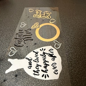 May include: Transparent sheet with wedding-themed designs. Includes a gold ring, a white dress silhouette, hearts, and the phrases "I said yes," "future Mrs," and "and they lived happily ever after" in black script.