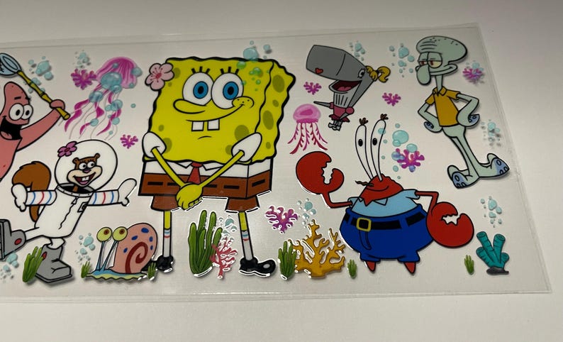 May include: A clear sticker sheet featuring cartoon characters from the animated series, including SpongeBob SquarePants, Patrick Star, Sandy Cheeks, Squidward Tentacles, Mr. Krabs, and Pearl. The characters are in bright colors with a cartoon style.