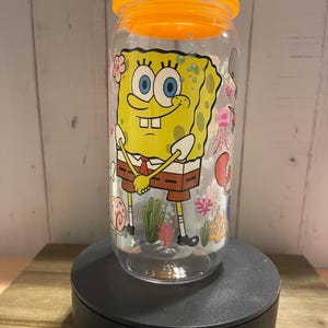 May include: Clear plastic tumbler with an orange lid featuring a cartoon image of SpongeBob SquarePants. The tumbler has a wraparound design with sea creatures and plants. The tumbler is on a black stand.