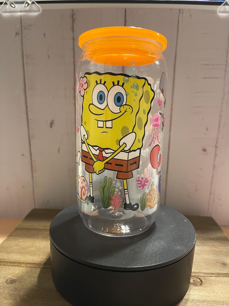 May include: Clear plastic tumbler with an orange lid featuring a cartoon image of SpongeBob SquarePants. The tumbler has a wraparound design with sea creatures and plants. The tumbler is on a black stand.