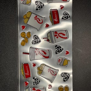 May include: A sheet of Chick-fil-A stickers featuring various food items, including chicken nuggets, waffle fries, chicken sandwiches, and drinks. The stickers are on a silver background with red hearts and black and white cow print hearts.