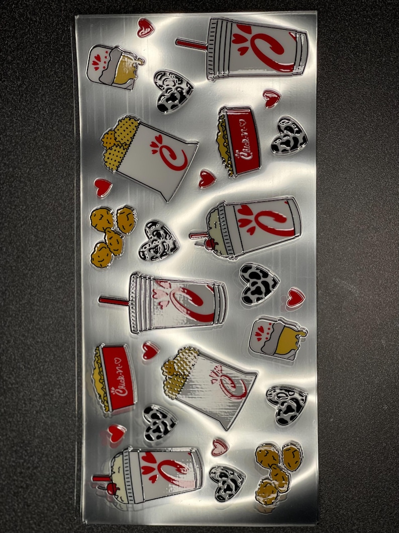 May include: A sheet of Chick-fil-A stickers featuring various food items, including chicken nuggets, waffle fries, chicken sandwiches, and drinks. The stickers are on a silver background with red hearts and black and white cow print hearts.