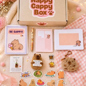 Puede incluir: Un set de regalo 'Happy Cappy Box' con varios art&iacute;culos tem&aacute;ticos de carpincho. Incluye una caja, tarjetas de felicitaci&oacute;n, pegatinas, un bol&iacute;grafo, un llavero y un peluche. La caja y las tarjetas muestran el texto 'Happy Cappy Box' y 'Be Cappy!'