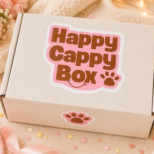 Puede incluir: Una caja de cart&oacute;n beige con una pegatina rosa y marr&oacute;n que dice "Happy Cappy Box" con una huella de pata. La caja est&aacute; sobre una superficie suave y texturizada con flores rosas y cinta en el fondo.