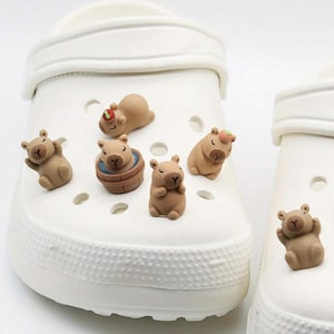 May include: A set of seven brown capybara charms, each with a different pose, including one in a wooden tub. The charms are designed to be worn on shoes or bags.