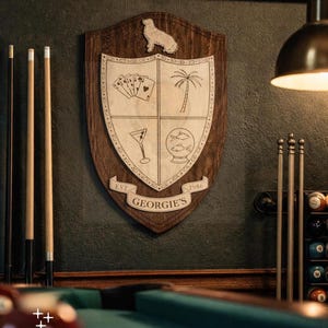 May include: A wooden shield-shaped plaque with a crest featuring a dog silhouette, playing cards, a palm tree, a martini glass, and fish in a snow globe. The plaque is mounted on a dark green wall, with pool cues and a pool table visible.