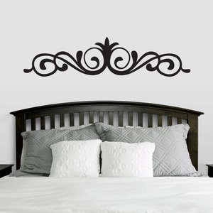 May include: Black decorative wall decal with a swirling design, above a dark brown wooden headboard with white and gray pillows on a white bed.