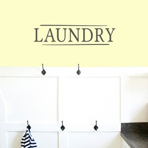 May include: A white wall with the word "LAUNDRY" written in gray lettering with two horizontal lines above and below the word. The wall is above a white cabinet with black hooks and a gray countertop.