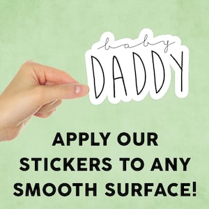 May include: A white sticker with black text that reads "baby daddy". A hand is holding the sticker up against a light green background. The text below the sticker reads "Apply our stickers to any smooth surface!"