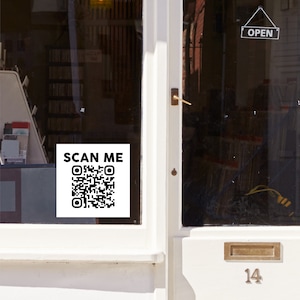 May include: A white sign with black text that says "SCAN ME" and a black and white QR code. The sign is attached to a glass door with an "OPEN" sign hanging above it.