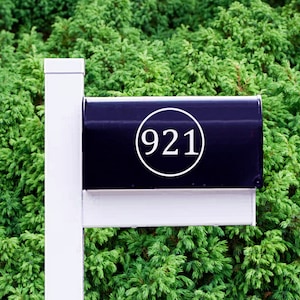 May include: Black mailbox with a white circle and the number 921 inside. The mailbox is mounted on a white post.