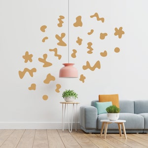 May include: A living room with a light grey sofa, a small wooden side table, and a white side table with a plant on top. The wall is white with a light brown abstract design. A pink pendant light hangs from the ceiling.