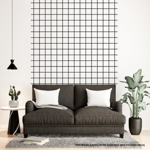 May include: A brown couch with white pillows sits in front of a white wall with a black grid pattern decal. A potted plant and a side table with a small potted plant are on either side of the couch.