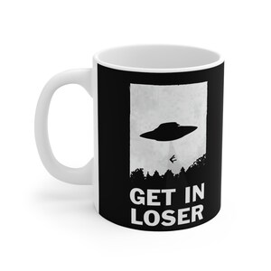 May include: White ceramic coffee mug with a black design. The design features a flying saucer over a forest with the text "GET IN LOSER" below.