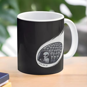 May include: A black and white ceramic coffee mug featuring a cartoon illustration of aliens in a vehicle. The illustration includes the text "ATLAS 3i". The mug has a white handle and interior.
