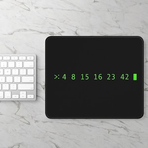 May include: Black rectangular mousepad with the numbers ">: 4 8 15 16 23 42" in green text. A white keyboard is visible in the upper left corner. The mousepad is on a white and gray marble-patterned surface.